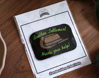 May include: Embroidered patch on a white card with a black border. The patch is black with green text that reads "Another Settlement Needs your help!" and a brown hat design. The card has a hat logo and the Instagram handle @embroidered.dad.hats.