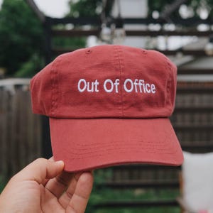 May include: A brick red baseball cap featuring the text Out Of Office embroidered in white thread across the front panel. The cap has a structured crown, a curved brim with visible stitching, and a button at the top center of the fabric dome.