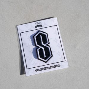 May include: A black and white embroidered patch in the shape of the letter "S". The patch is on a white card with a hat graphic and the text "@embroidered.dad.hats".