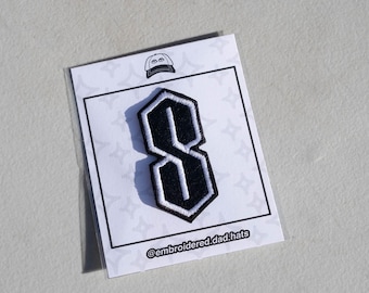 May include: A black and white embroidered patch in the shape of the letter "S". The patch is on a white card with a hat graphic and the text "@embroidered.dad.hats".