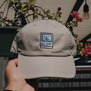 Può includere: Un cappello da baseball beige con un'immagine ricamata di un computer Macintosh vintage con un viso sorridente.