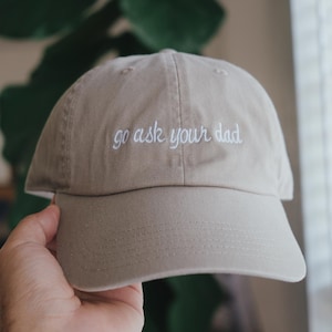 May include: A beige baseball cap with the text "go ask your dad" embroidered in white on the front.