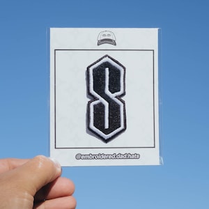 May include: A black and white embroidered patch featuring the iconic "S" design. The patch is in a clear plastic sleeve against a blue sky background. The text "@embroidered.dad.hats" is visible.