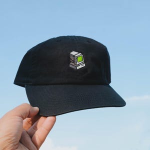 May include: A black baseball cap featuring a small, centered embroidered graphic of a vintage desktop computer. The computer is depicted in shades of grey with a bright, pixelated lime green screen, showcasing a classic retro aesthetic design.