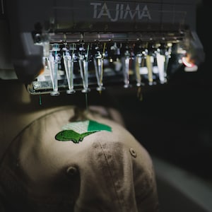 May include: An embroidery machine stitching a green and white design onto a tan baseball cap. The machine, labeled "TAJIMA," has multiple needles and threads. The cap features a small, embroidered graphic.