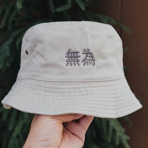May include: A beige bucket hat with the embroidered characters "無為" in a darker beige thread. The hat has a classic brim and is made of a soft, textured fabric. The hat is a neutral color and suitable for various outfits.