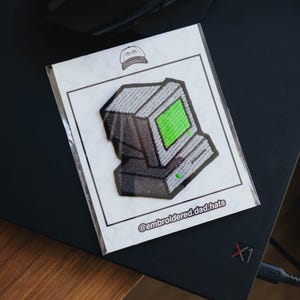 Peut inclure: Écusson brodé représentant un ordinateur rétro avec un écran vert. Le patch est gris et noir, avec le texte "@embroidered.dad.hats" en bas. L'écusson est sur une carte blanche.