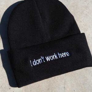 Puede incluir: Un gorro de punto negro con un dobladillo plegado que presenta el texto bordado en blanco "I don't work here." El gorro está confeccionado con un tejido negro sólido con una textura acanalada visible y una forma redondeada clásica.