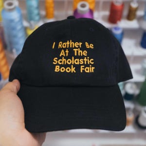 May include: A black baseball cap featuring yellow embroidered text that reads I Rather Be At The Scholastic Book Fair. The cap has a structured crown, a curved brim, and a button at the top center, showcasing a clean and simple design.