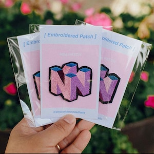 May include: An embroidered patch featuring a stylized N64 logo in shades of pink, purple, and blue with a black border. The patch is mounted on a white card with the text [ Embroidered Patch ] and @embroidereddadhats in clear plastic.