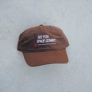 Peut inclure: Casquette de baseball marron avec une visière incurvée. Le texte brodé en blanc sur le devant dit "SEE YOU SPACE COWBOY..." avec un petit avion rouge en dessous. La casquette est en matière souple.