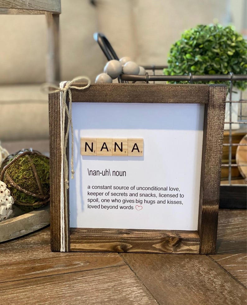 May include: A framed print with the word "NANA" spelled out in Scrabble tiles. The print defines "NANA" as a noun, a constant source of unconditional love, keeper of secrets and snacks, licensed to spoil, one who gives big hugs and kisses, loved beyond words.