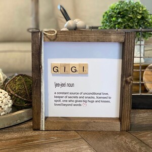 Pode incluir: Uma placa enquadrada em madeira com a palavra "GIGI" escrita em pe&ccedil;as de Scrabble. A placa define "GIGI" como um "nome jee-jeel" e descreve-a como uma fonte constante de amor incondicional, guardi&atilde; de segredos e lanches, autorizada a mimar, algu&eacute;m que d&aacute; grandes abra&ccedil;os e beijos, amada al&eacute;m das palavras.