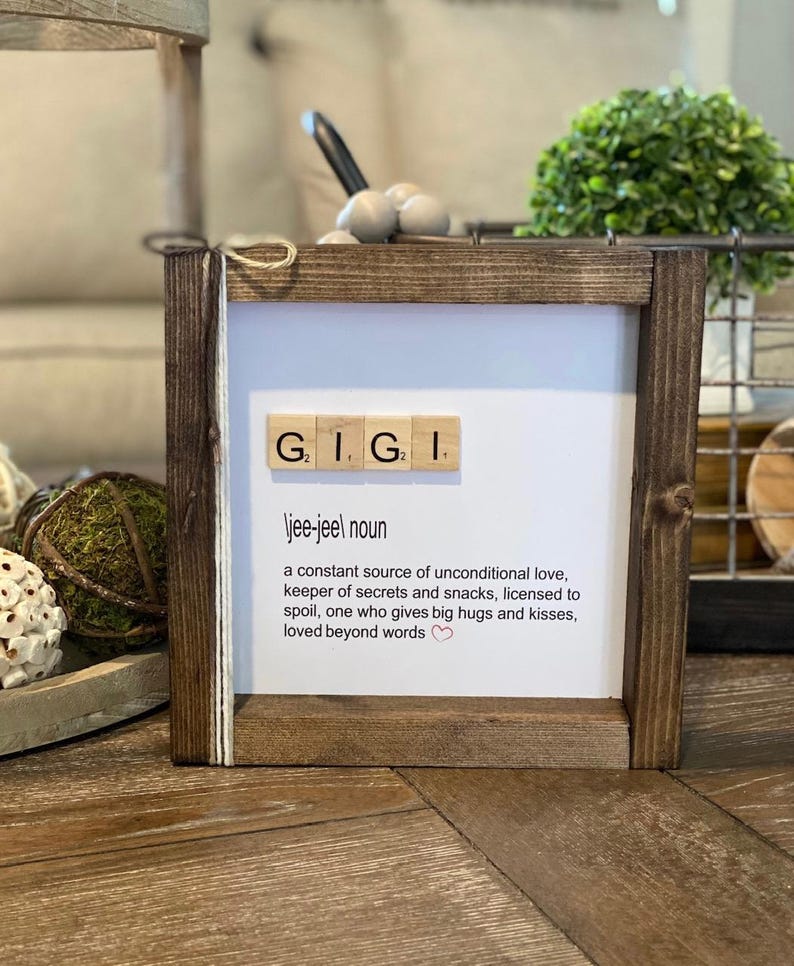Pode incluir: Uma placa enquadrada em madeira com a palavra "GIGI" escrita em pe&ccedil;as de Scrabble. A placa define "GIGI" como um "nome jee-jeel" e descreve-a como uma fonte constante de amor incondicional, guardi&atilde; de segredos e lanches, autorizada a mimar, algu&eacute;m que d&aacute; grandes abra&ccedil;os e beijos, amada al&eacute;m das palavras.