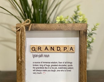 May include: A wooden framed sign with the word "GRANDPA" spelled out in Scrabble tiles. The sign also includes the definition of "grandpa" as "a source of immense wisdom, fixer of all things broken, king of hugs, greatest storyteller, spoils the grandkids like it is his job, indefinitely patient, will always make you laugh, one who is loved very much."