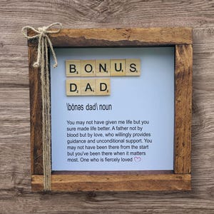 May include: A wooden framed sign with the words "BONUS DAD" spelled out with Scrabble tiles. The sign includes a definition and a heartfelt message about a father figure, with a twine bow on the side.