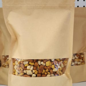 May include: A light brown kraft paper stand-up pouch with a clear rectangular window in the center. The pouch is filled with small, cubed pieces of dried food in shades of yellow, orange, and white, visible through the transparent plastic section.