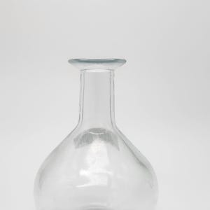 May include: A clear glass vase with a rounded body and a long, narrow neck. The vase has a wide, flat rim and sits on a clear base. The glass is transparent, allowing light to pass through. The vase is empty and sits against a white background.