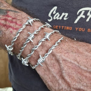 Könnte beinhalten: Vier silberne Stacheldrahtarmbänder, die auf dem Handgelenk einer Person übereinander liegen. Die Armbänder bestehen aus verdrehtem Draht mit scharfen Spitzen.