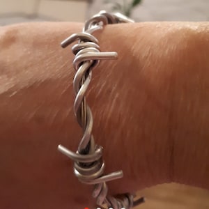 Puede incluir: Un brazalete de alambre de púas plateado. El brazalete está hecho de alambre retorcido con puntas afiladas.