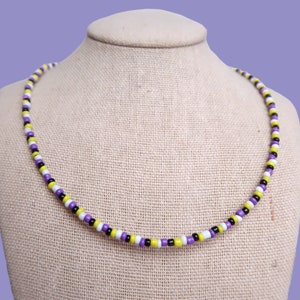 May include: A beaded necklace with alternating purple, white, yellow, and black beads. The necklace is on a fabric-covered mannequin.