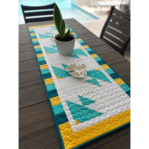 Könnte beinhalten: Ein gequilteter Tischläufer in den Farben Türkis, Gelb und Weiß mit einem geometrischen Muster. Der Läufer liegt auf einem Holztisch mit einer Topfpflanze im Hintergrund.