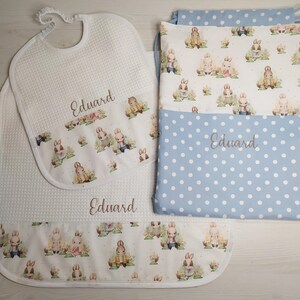 May include: A baby bedding set comprising a white waffle-weave bib, blanket, and pillowcase. The set features a bunny pattern and a blue polka dot design on the pillowcase. The name "Eduard" is embroidered on the items. The items are designed for a baby.