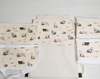 May include: A set of baby accessories featuring a cream-colored fabric with a repeating pattern of construction vehicles including dump trucks, excavators, and forklifts, consisting of two bibs, a drawstring backpack, and two folded white towels.