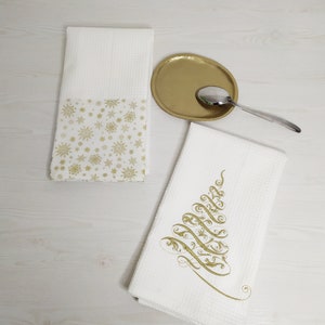 Puede incluir: Dos toallas de cocina blancas con bordados dorados. Una toalla presenta un patrón de copos de nieve, la otra tiene un diseño estilizado de árbol de Navidad.