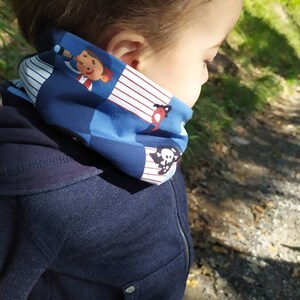 May include: A child wearing a blue hooded sweatshirt with a colorful neck gaiter featuring a pirate, a skull and crossbones, and other nautical designs.