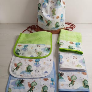 May include: A drawstring backpack and several towels featuring a repeating pattern of green dragons, planets, stars, and clouds on a white background. The towels have lime green or light blue trim and textured fabric sections in matching colours.