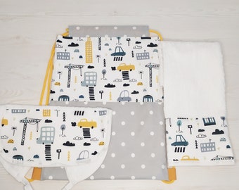 May include: A set of baby items featuring a white background with a repeating pattern of yellow cars, blue buildings, and gray clouds. The set includes a gray and white polka dot drawstring bag, a white bib with a yellow trim, and a white towel with a gray border.