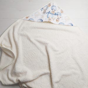 Puede incluir: Una toalla con capucha blanca para bebé. La capucha tiene un estampado de ositos de peluche en globos aerostáticos, nubes y el nombre "Amedeo" en escritura azul. La toalla está hecha de material suave y absorbente.