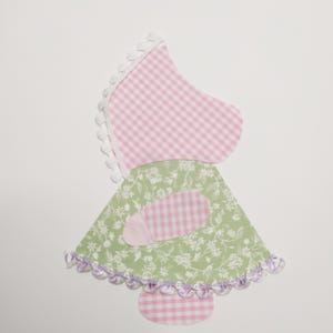 May include: A pink and green fabric doll with a white trim and a pink gingham dress. The doll has a green skirt with white flowers and a pink gingham bow.