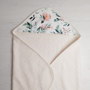 Puede incluir: Toalla de bebé con capucha de color crema con una capucha con temática de bosque. La capucha presenta un patrón de ardilla, conejo y ciervo entre hojas verdes y el nombre "Bianca" en escritura rosa. La toalla está hecha de material suave y absorbente.
