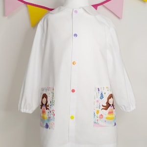 Puede incluir: Una bata blanca de manga larga con cuello redondo y cierre frontal de botones multicolores. La prenda incluye dos bolsillos delanteros, cada uno decorado con una colorida ilustración de una niña con vestido rodeada de flores.