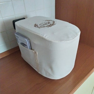 Könnte beinhalten: Weiße Stoffhülle für ein Küchengerät mit einem braunen Stickmuster und einer Tasche an der Seite.