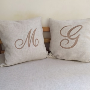 Op de afbeelding: Twee beige linnen kussens met geborduurde cursieve letters, een met een "M" en de andere met een "G".