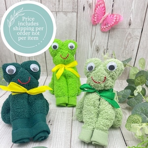 Puede incluir: Tres ranas de toalla hechas a mano en tonos verdes, cada una con ojos saltones y un lazo de cinta. Una rana es verde oscuro, otra verde brillante y otra verde claro. Una mariposa rosa y texto en el fondo.
