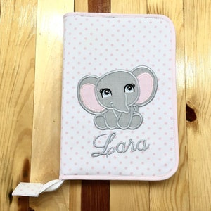 Può includere: Un libro per bambini in tessuto a pois rosa e bianco con un elefante ricamato grigio e il nome "Lara" sulla copertina.