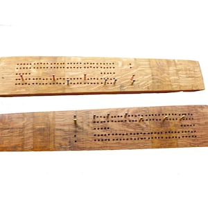 May include: A wooden cribbage board made from a repurposed barrel stave. The board has two sections, each with a grid of holes for pegs. The board is stained a light brown colour and has a natural wood grain.