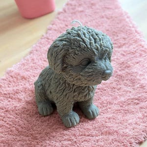 May include: A grey, furry, dog-shaped candle with a white wick. The candle is sitting on a pink, fluffy surface.