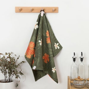 May include: A green kitchen towel with a floral pattern of orange and white hibiscus flowers and green leaves. The towel is hanging on a wooden peg rack against a white wall. The towel is made of a waffle weave fabric.