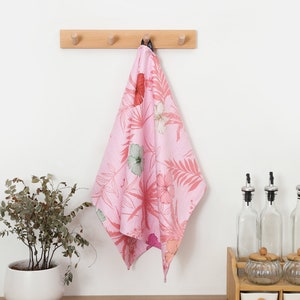 May include: A pink kitchen towel with a floral print hangs on a wooden hook rack. The towel features a pattern of pink hibiscus flowers and green leaves.