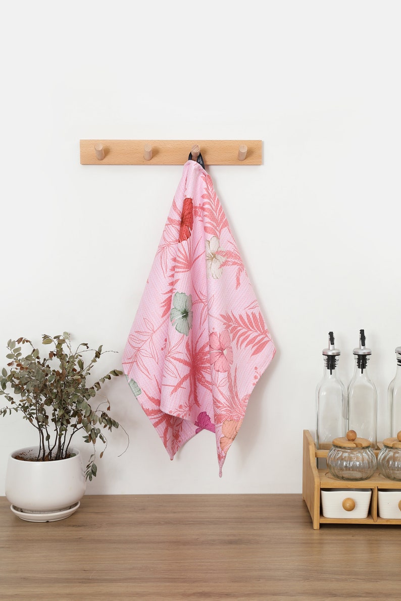 May include: A pink kitchen towel with a floral print hangs on a wooden hook rack. The towel features a pattern of pink hibiscus flowers and green leaves.