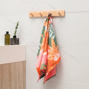 K&ouml;nnte beinhalten: Ein bunt gemustertes Handtuch h&auml;ngt an einer Holzleiste mit f&uuml;nf Kn&ouml;pfen an einer wei&szlig; gefliesten Wand. Das Handtuch hat ein lebendiges Design mit orangefarbenen, gr&uuml;nen und rosa Blumen- und Bl&auml;ttermustern. Badezimmerdekoration.