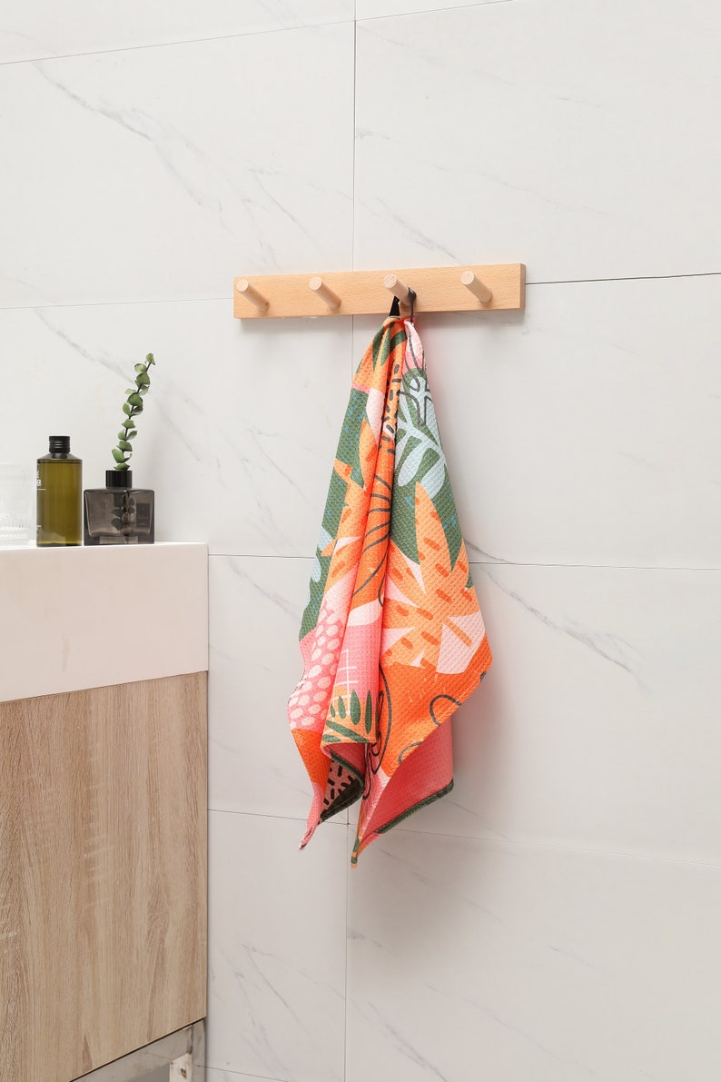 K&ouml;nnte beinhalten: Ein bunt gemustertes Handtuch h&auml;ngt an einer Holzleiste mit f&uuml;nf Kn&ouml;pfen an einer wei&szlig; gefliesten Wand. Das Handtuch hat ein lebendiges Design mit orangefarbenen, gr&uuml;nen und rosa Blumen- und Bl&auml;ttermustern. Badezimmerdekoration.