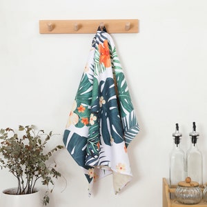 May include: A white kitchen towel with a tropical floral pattern of green leaves and orange and yellow flowers hangs from a wooden peg rack. The towel is made of a textured fabric and is hanging in a kitchen setting.