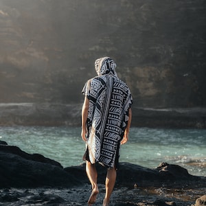 May include: A person wearing a black and white hooded poncho with a geometric pattern stands on a rocky beach. The person is facing away from the camera and looking out at the ocean.