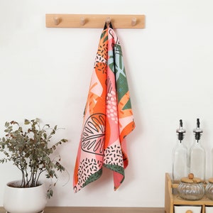 K&ouml;nnte beinhalten: Ein farbenfrohes K&uuml;chentuch mit einem lebendigen Zitrusfrucht-Design, das an einer Holzleiste h&auml;ngt. Das Tuch zeigt orange, rosa, gr&uuml;ne und schwarze Muster. Ein Blumentopf und K&uuml;chenzubeh&ouml;r befinden sich im Hintergrund.